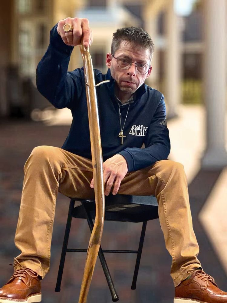 A man with glasses, short hair, wearing a dark jacket and tan pants, sitting on a black chair, holding a wooden cane upright, with a serious expression on his face.