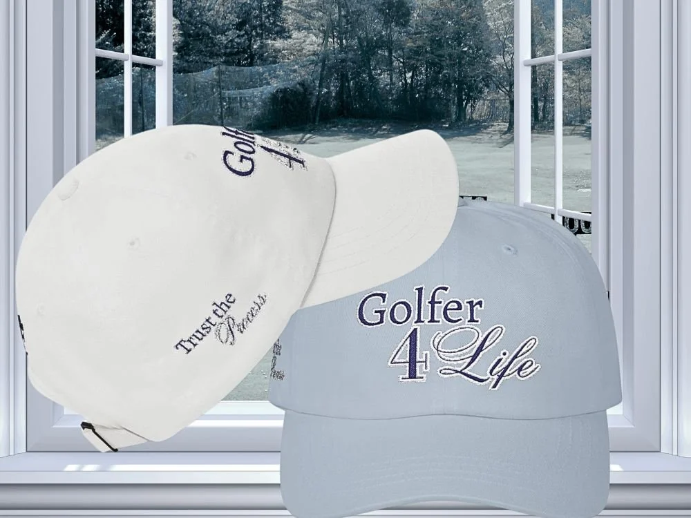 Two golf caps, one white and one light gray, with embroidered text, on a window ledge with a view of a golf course in the background.