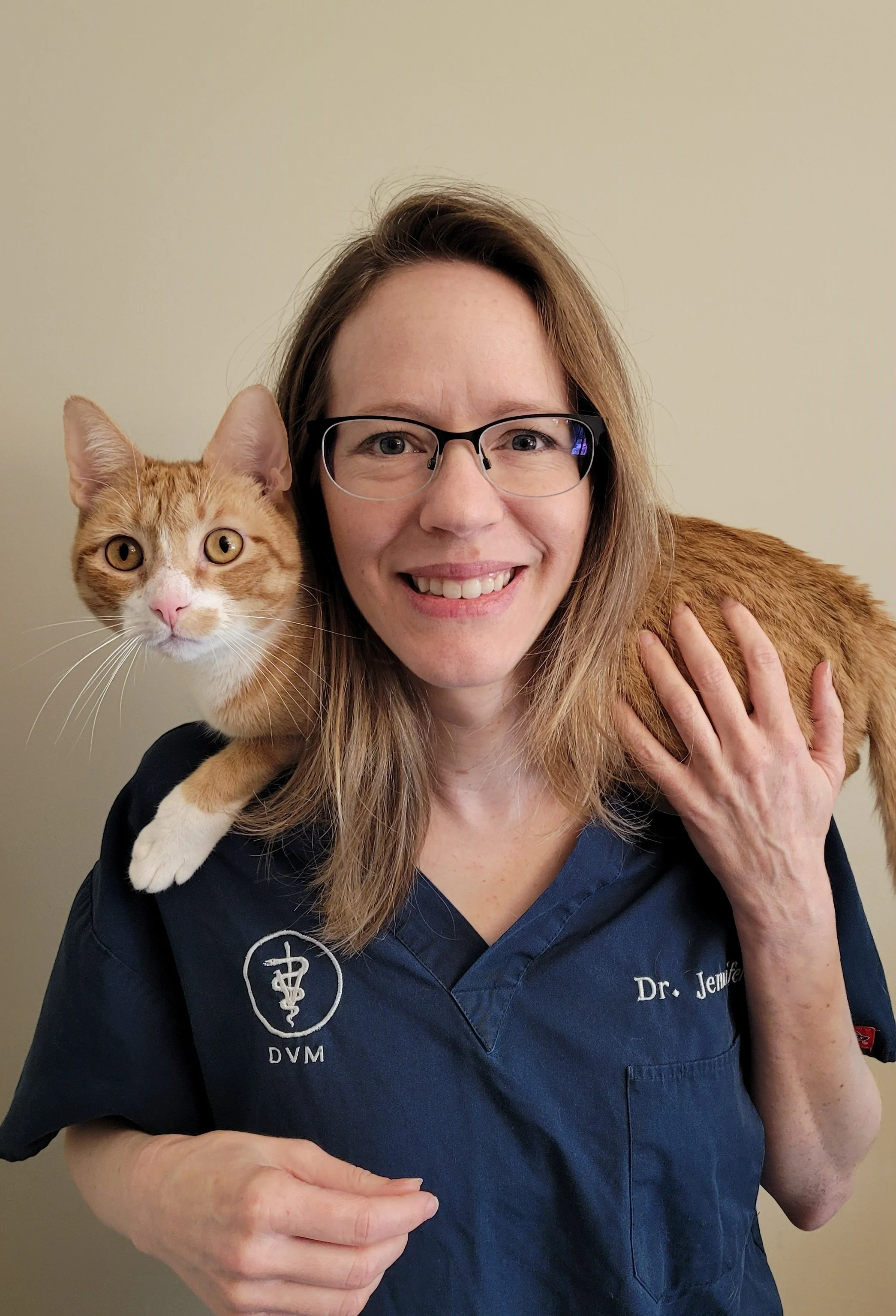 A woman with glasses wearing a navy veterinary coat, holding an orange tabby cat on her shoulder. The woman is smiling and has long, light brown hair. The cat has large yellow eyes and appears calm.