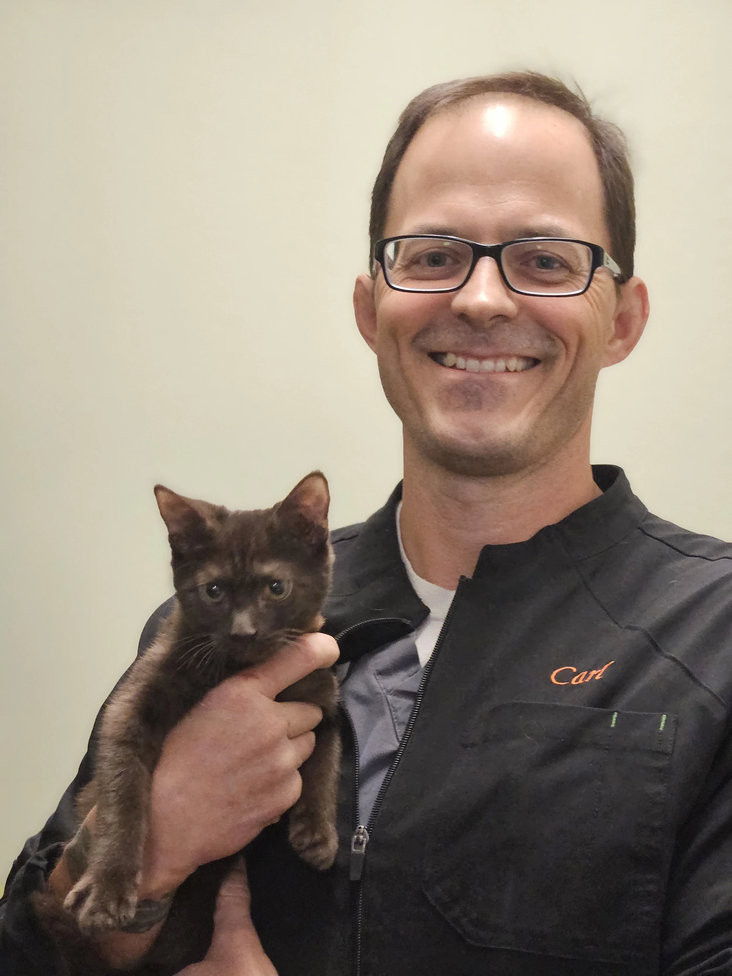 A smiling man with short brown hair, glasses, and a black jacket with the name 'Carl' embroidered on it, holding a small brown and black kitten.