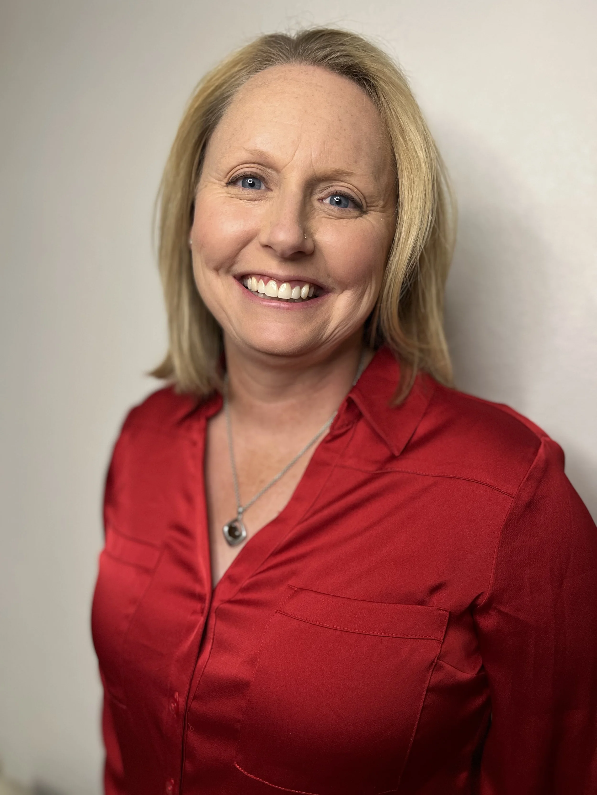 A woman with blonde hair smiling, wearing a red blouse and a silver necklace, standing against a plain wall.