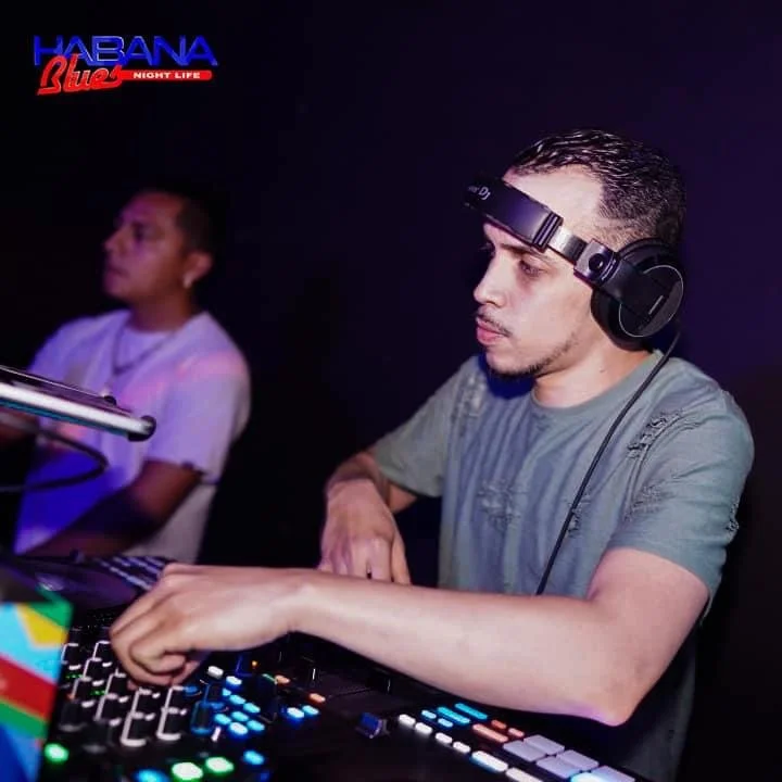 A DJ wearing headphones is mixing music at a nightclub with colorful lights. Another person stands in the background. The logo in the top left corner reads "Habana Club Night Life."