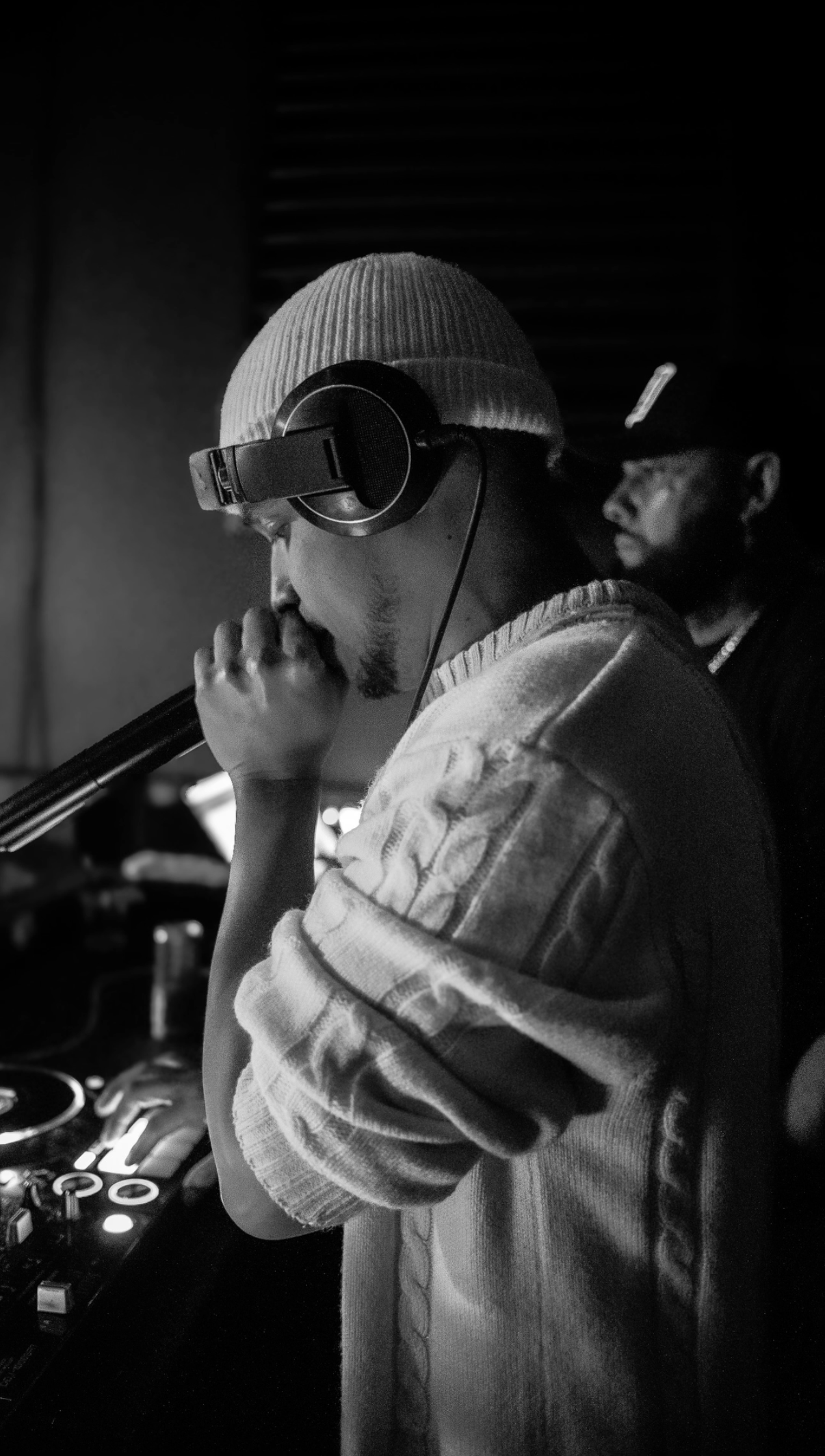 A black and white photo of a DJ wearing headphones, a beanie, and a sweater, standing in front of a DJ mixer in a dark room.