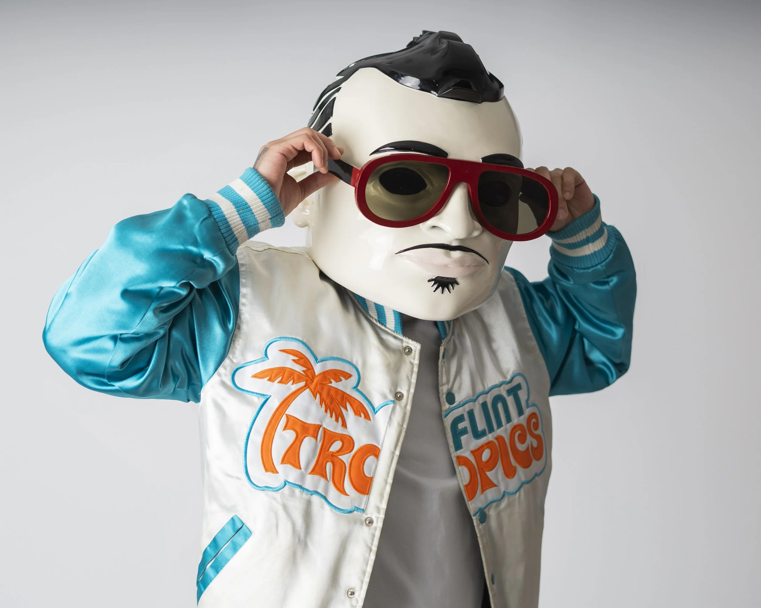 Person wearing a retro-style varsity jacket with 'Flint Tropics' logo, red sunglasses, and a mannequin mask with black and white hair.