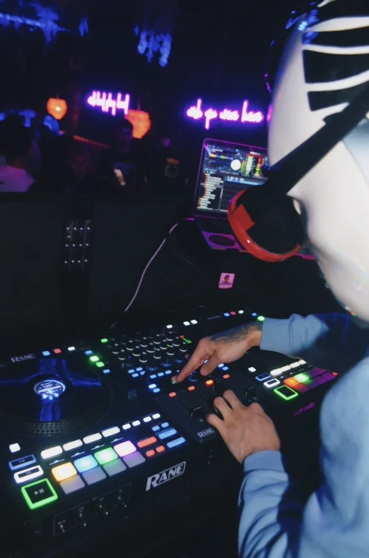 DJ wearing a helmet with red headphones, operating a DJ controller with colorful lights in a dark club environment, with a laptop displaying DJ software in the background.