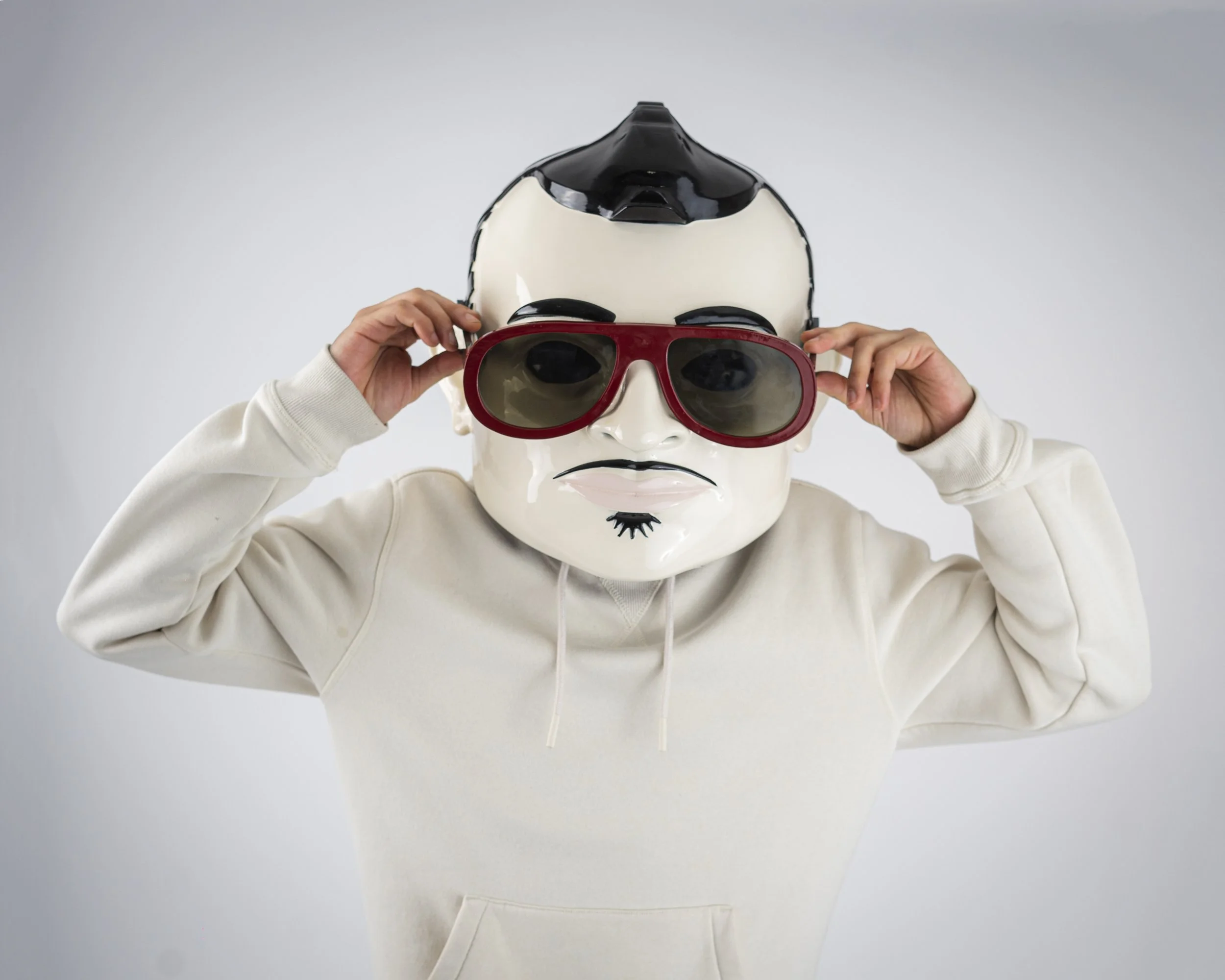 Person is wearing a mask resembling a stylized face with exaggerated features, and adjusting large red sunglasses. They are dressed in a cream-colored hoodie.