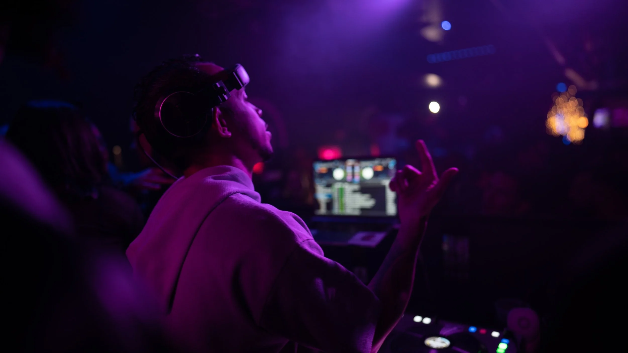 A DJ in a dimly lit club wearing headphones, pointing with one hand, with colorful lights and DJ equipment in the background.