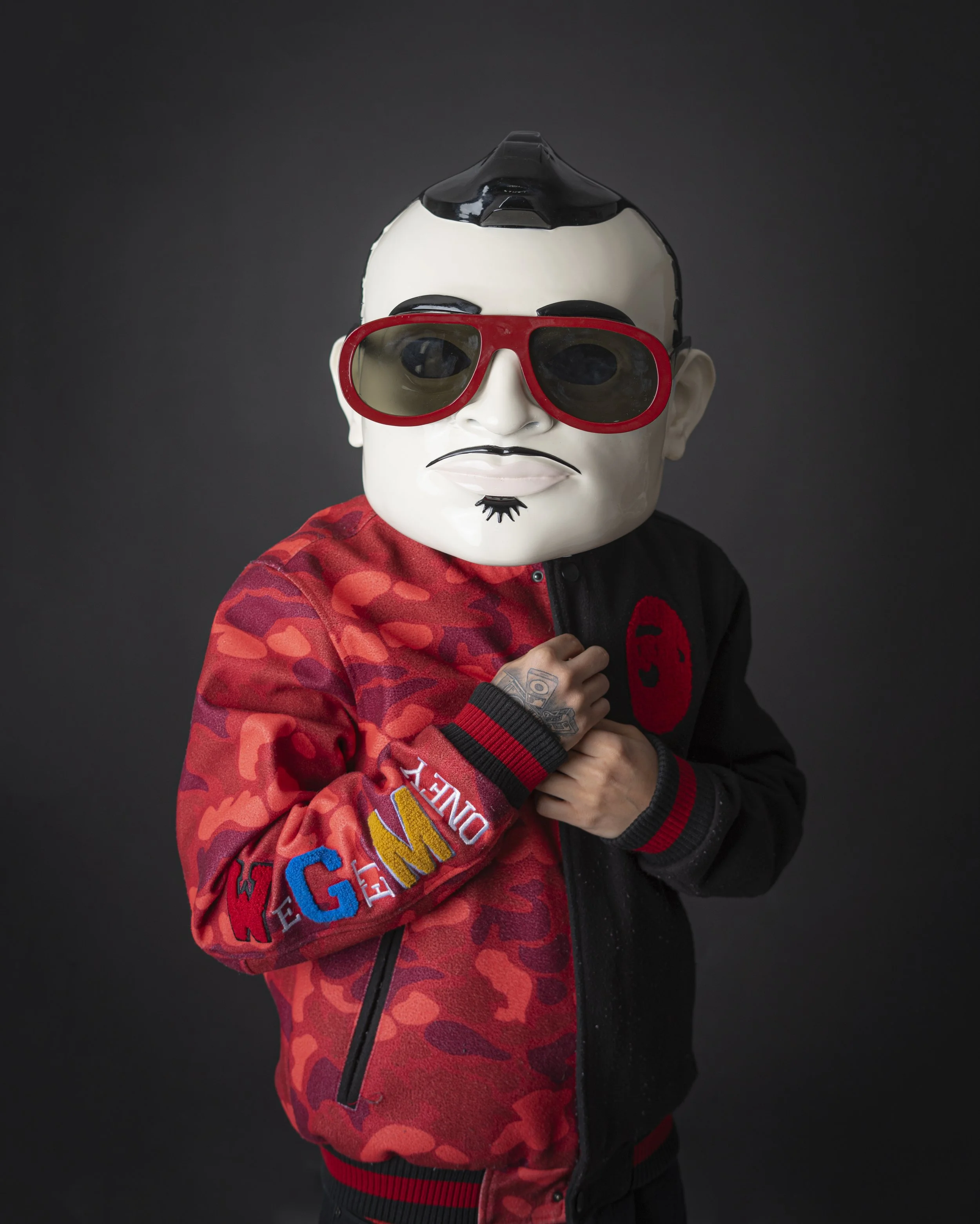 Person wearing a large cartoonish mask with sunglasses, a red and black jacket, and standing against a dark background.