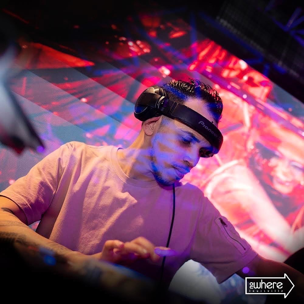 A DJ wearing headphones and a brown t-shirt, mixing music on a DJ controller in a nightclub with colorful LED lights.