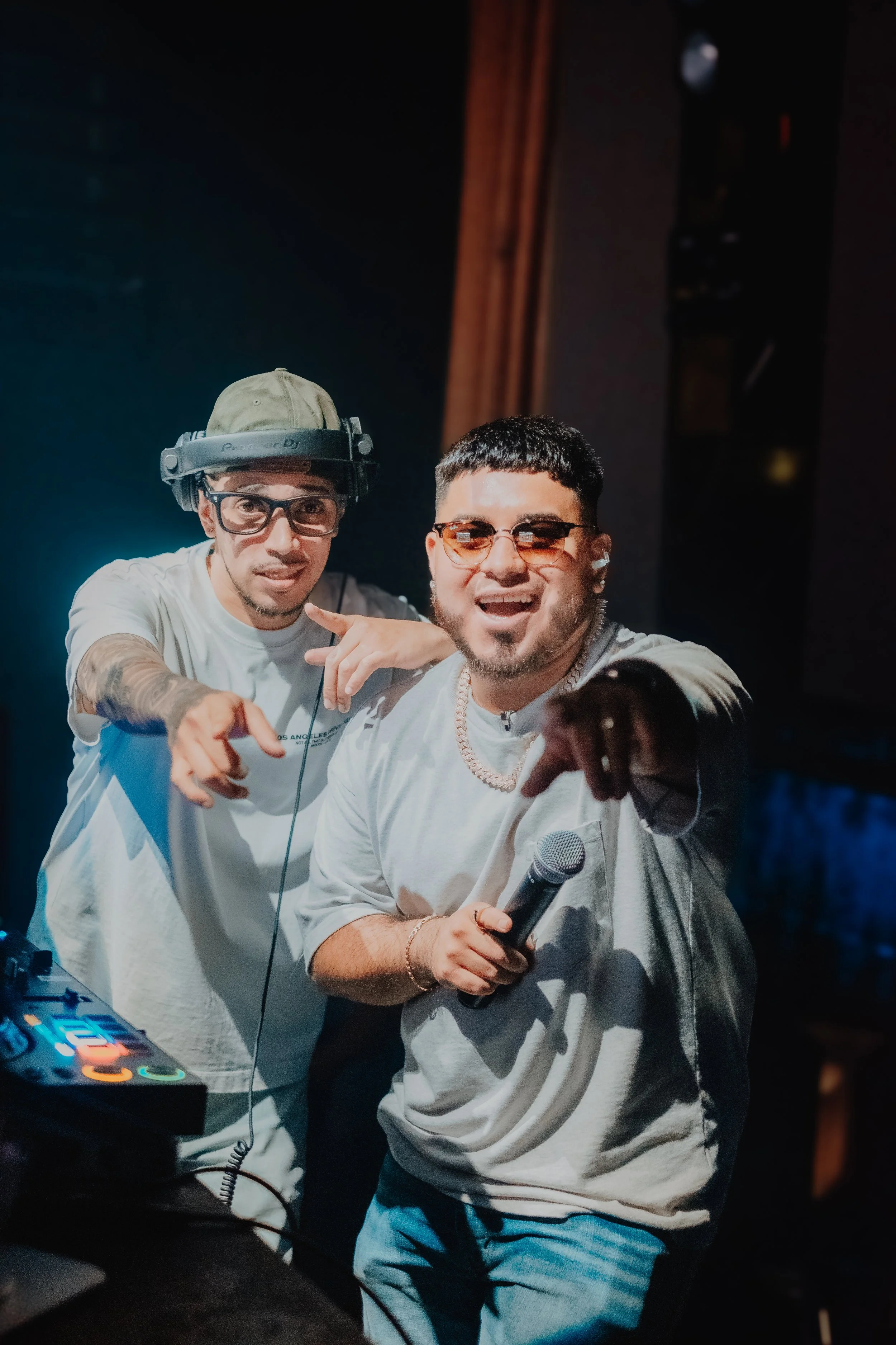 Two men in a nightclub, one wearing a cap and headphones and the other with sunglasses and a microphone, posing together while DJ equipment is visible.