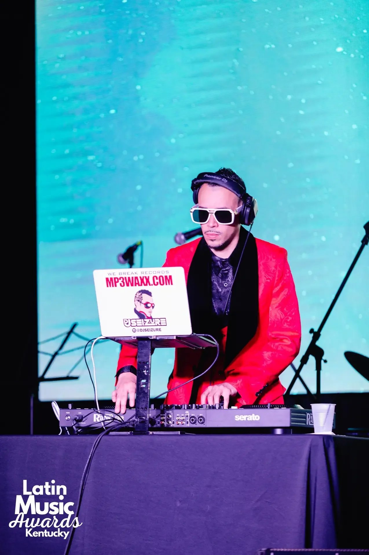 DJ in red blazer, sunglasses, headphones, standing at DJ equipment, with a blue digital background, at Latin Music Awards Kentucky.