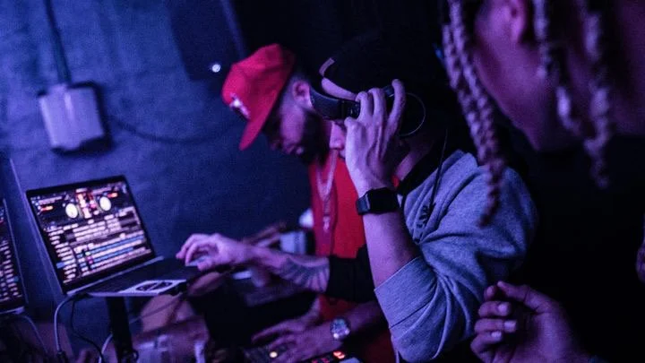 A DJ wearing a red cap and hoodie using turntables and a laptop at a club or party setting.