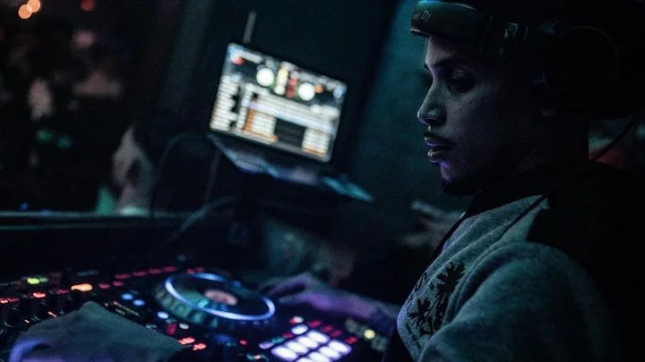 A DJ wearing headphones, operating DJ equipment in a dark environment with a computer screen in the background.