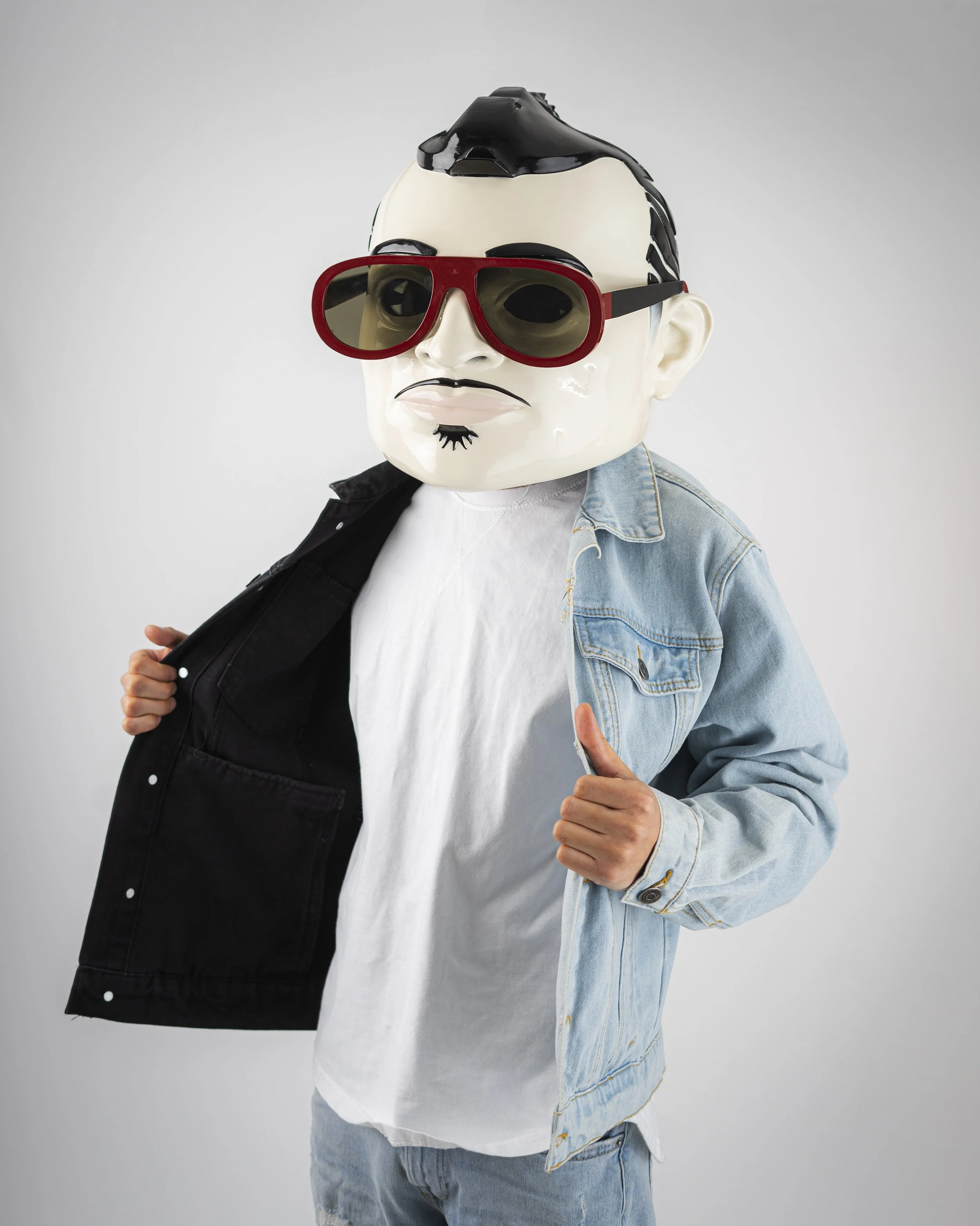 Person wearing a ceramic mask with a stylized face, large red sunglasses, a white shirt, a denim jacket, and light blue jeans, giving a thumbs-up.