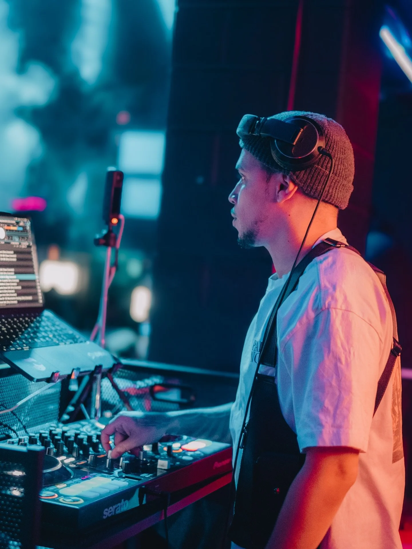A DJ wearing a gray beanie and headphones is standing at a mixing console, operating electronic music equipment in a nightclub with colorful lighting.