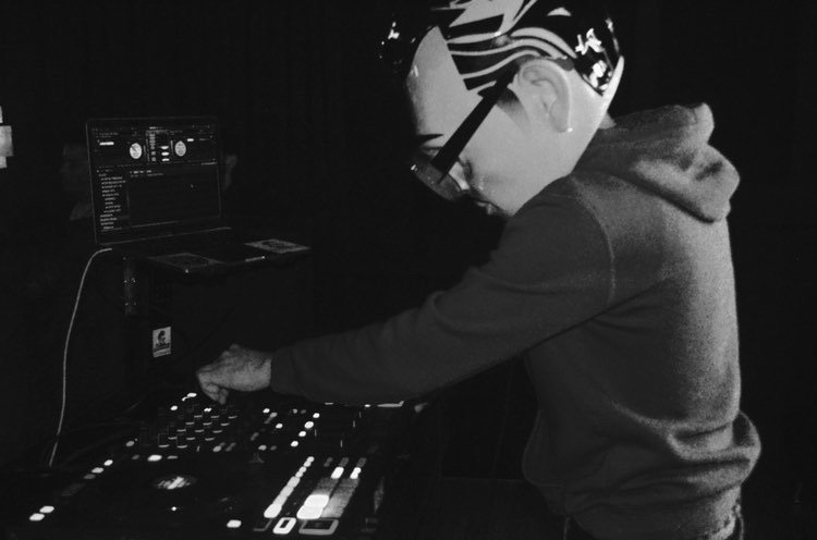 Person DJing with equipment in a dark setting, wearing glasses and a hoodie.
