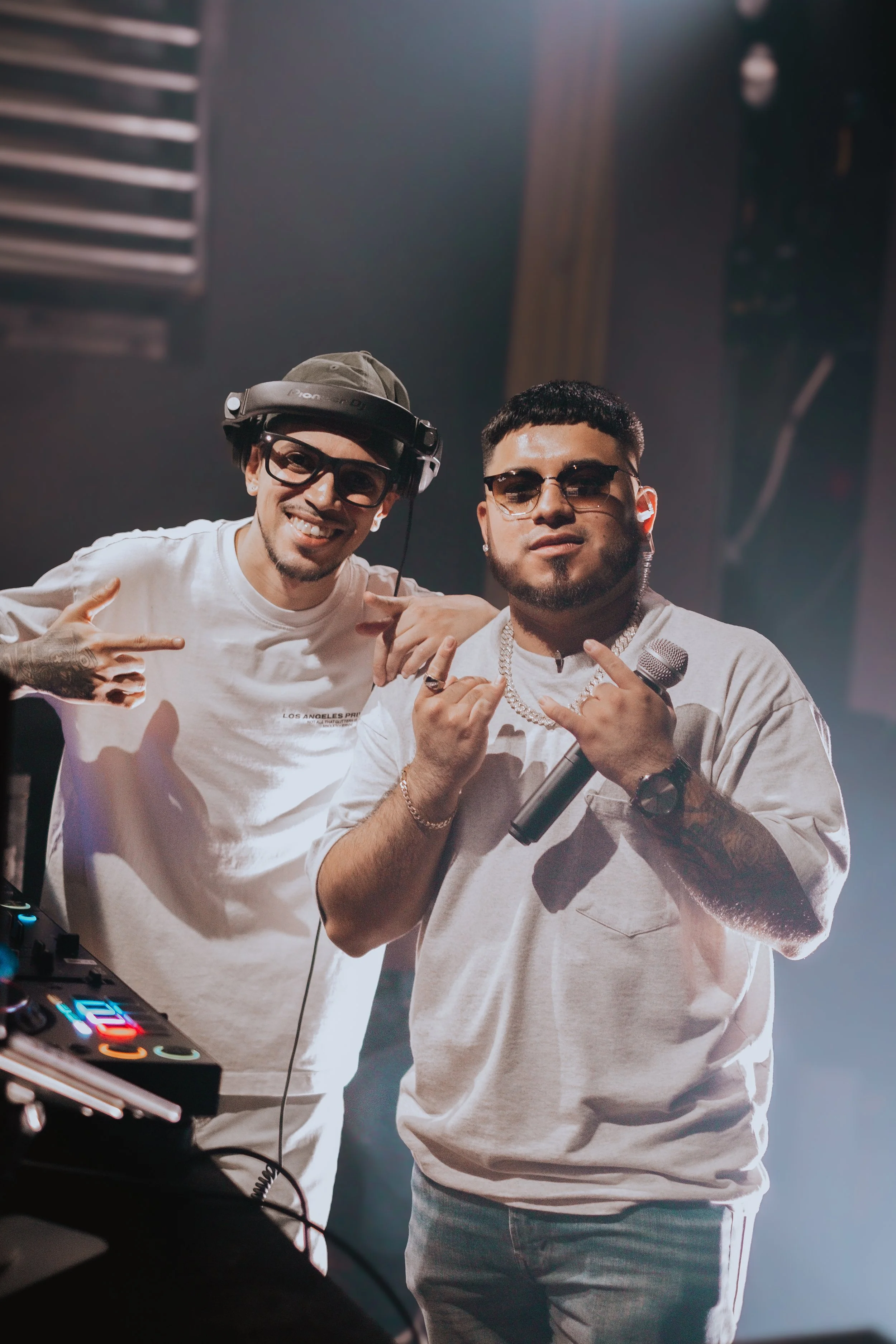 Two men smiling and posing backstage at a music event, one wearing headphones and the other holding a microphone, with DJ equipment visible in the foreground.