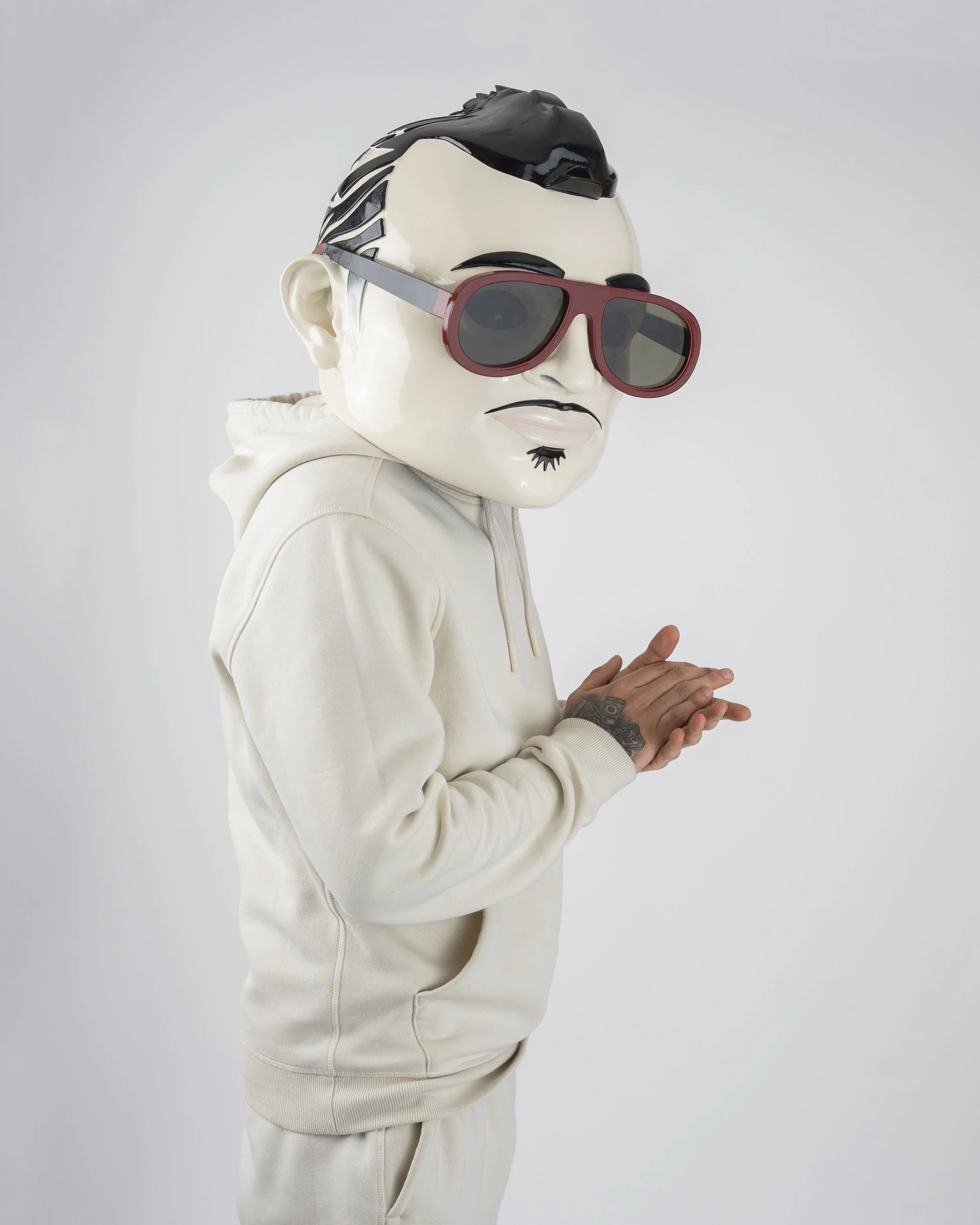 Person wearing a large cartoonish mask with sunglasses and hooded sweatshirt, hands clasped together.