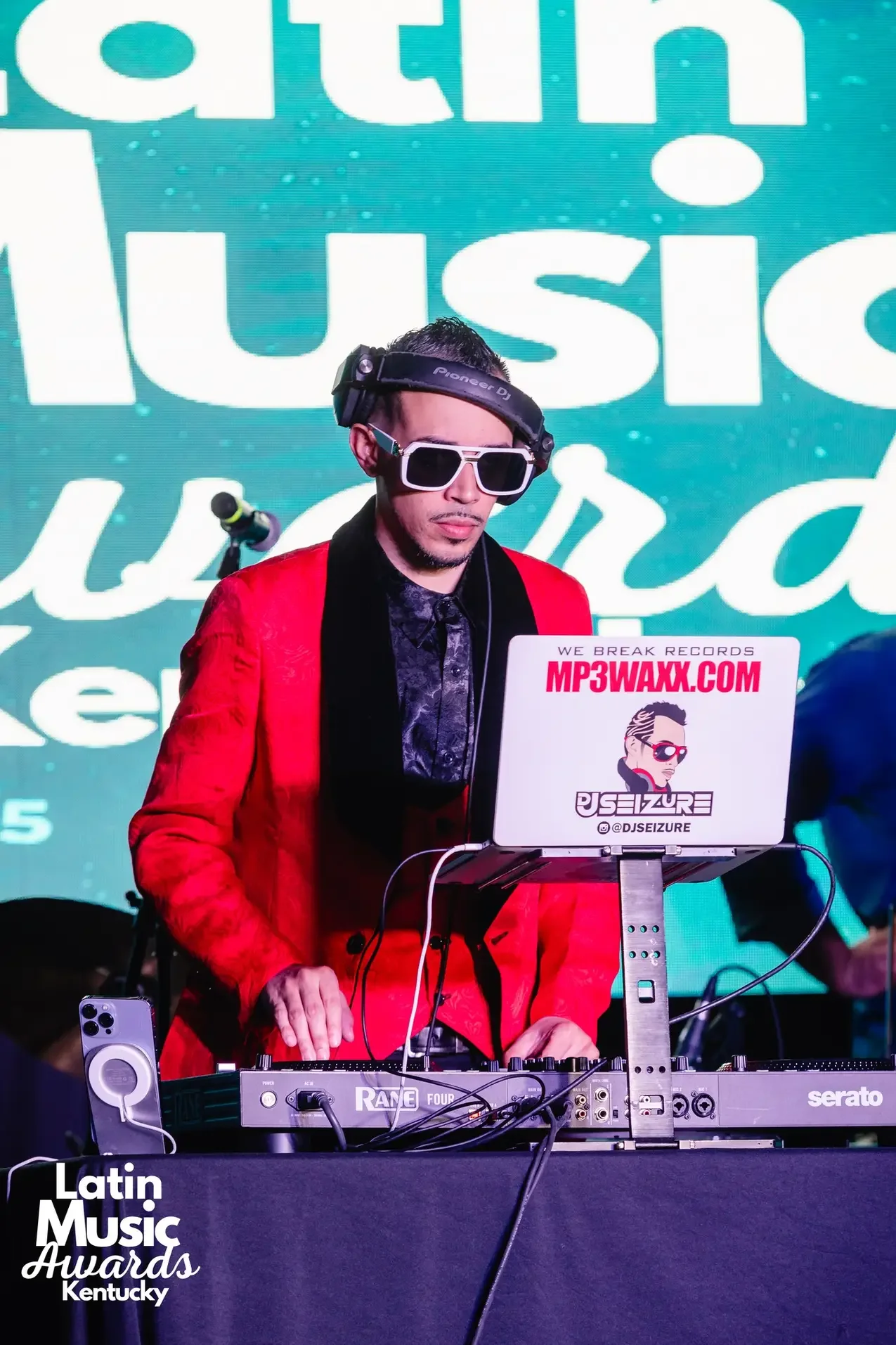 A DJ wearing a red blazer, black shirt, sunglasses, and headphones is performing at a music event. He is standing behind a DJ console with a laptop titled 'MP3WAXX.COM' and a sticker that says '@DJSEIZURE'. In the background, there is a blue screen w