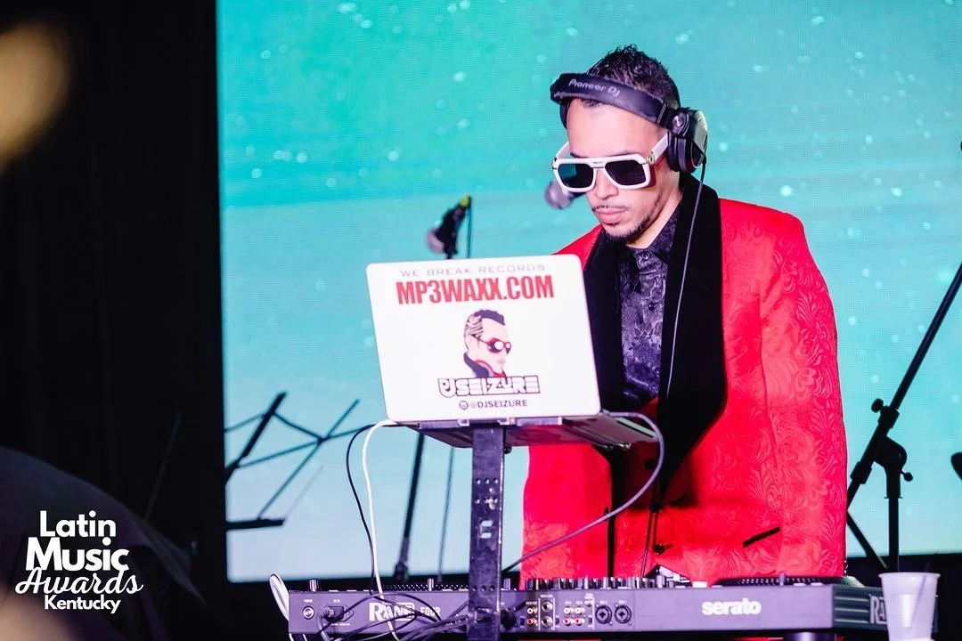 DJ wearing white sunglasses, headphones, and a red jacket, performing with a DJ controller on stage at the Latin Music Awards in Kentucky.