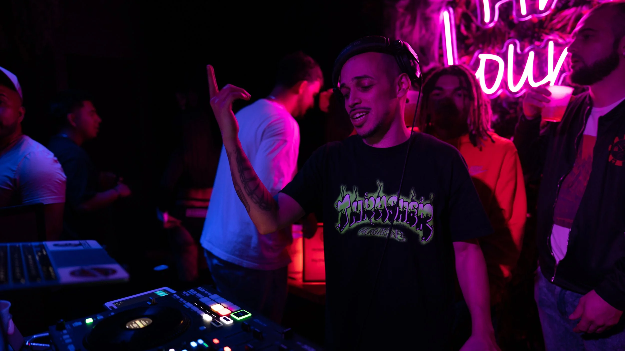 A DJ with headphones and a Thrasher magazine t-shirt performs on his DJ equipment while dancing at a nightclub, surrounded by people with neon signs in the background.