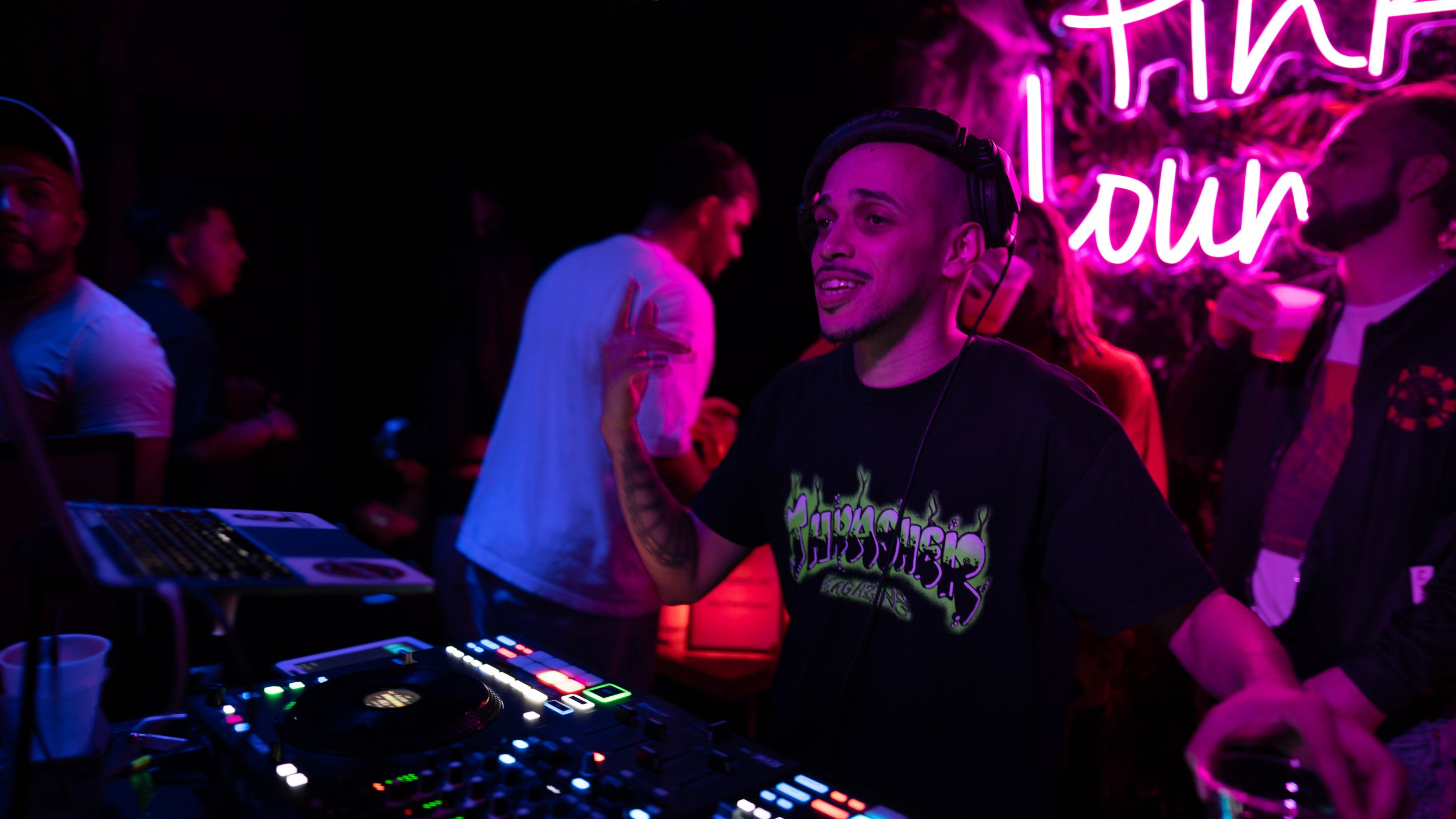 A DJ is mixing music at a nightclub, surrounded by people dancing and socializing, with neon pink and purple lights and a neon sign reading 'that' and 'our' in the background.