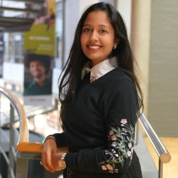 Een jonge vrouw met lang zwart haar en een witte blouse, gekleed in een zwarte jas met bloemmotief, glimlachend naar de camera, binnen in een gebouw met ramen en posters op de achtergrond.