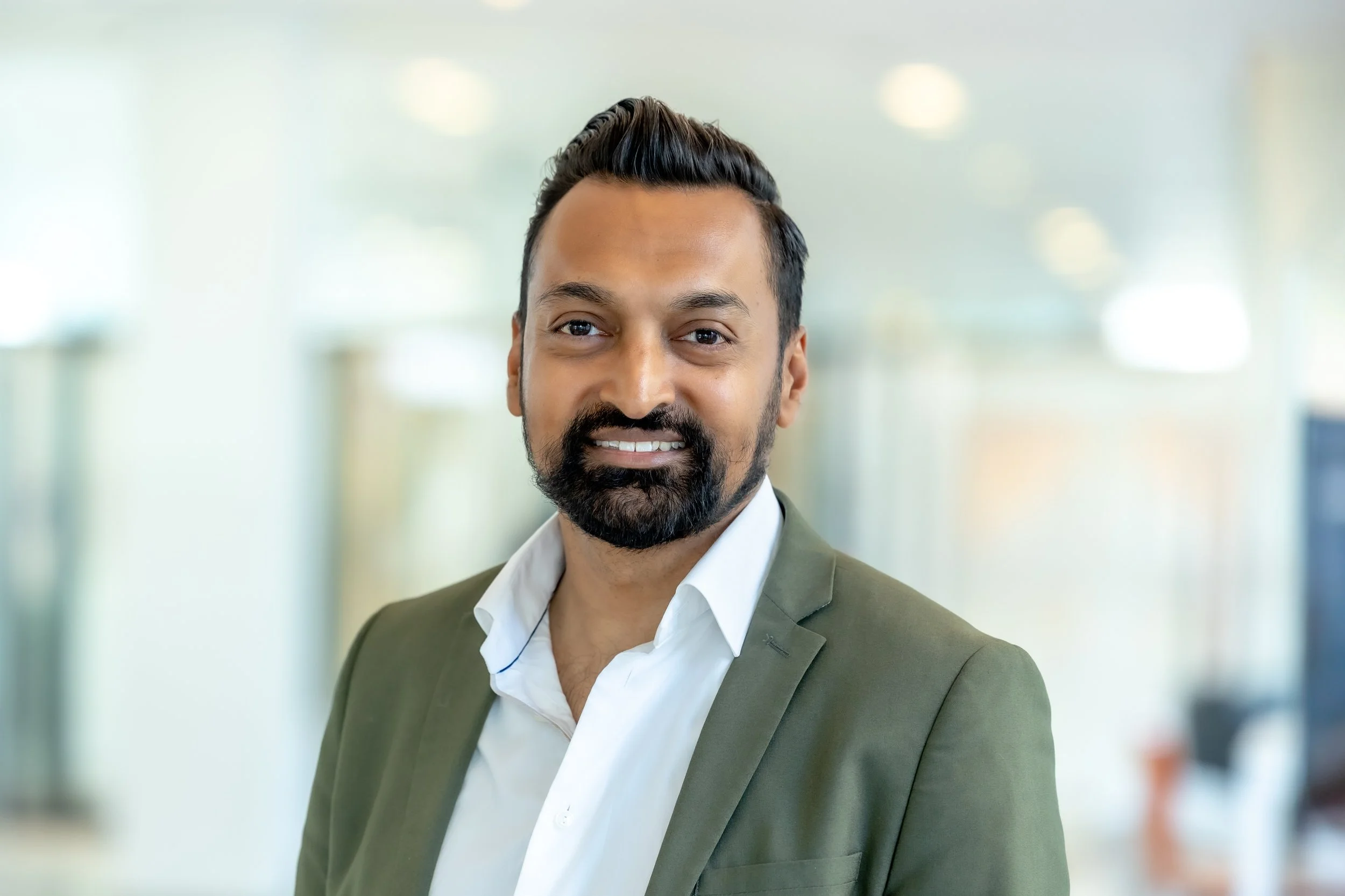 Persoon met donkere haar en baard, lachend, in een groen jasje en witte shirt, binnen in een kantoor.