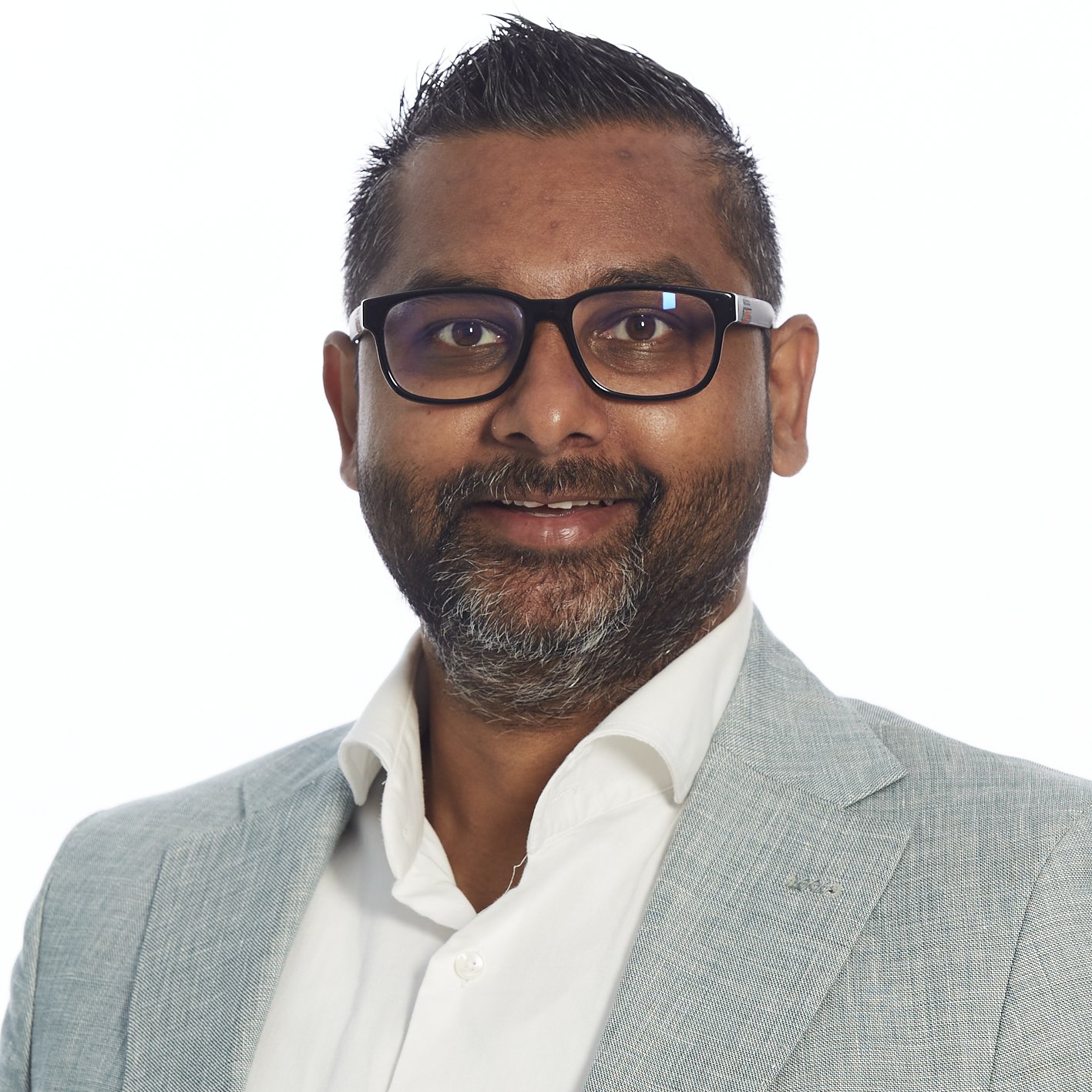 Professioneel portret van een man met een grijze jas, wit overhemd en zwarte bril, lachend tegen een witte achtergrond.