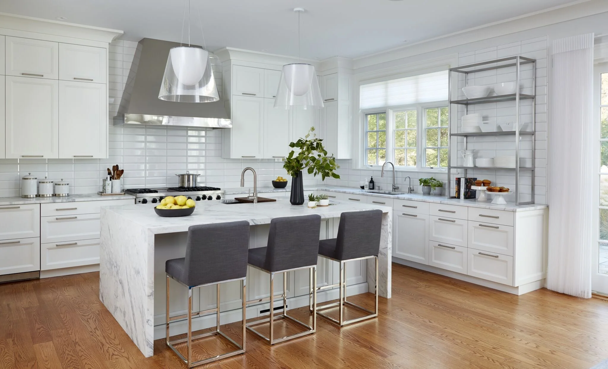 Modern white kitchen with marble island, gray barstools, stainless steel appliances, open shelving, and large windows letting in natural light.