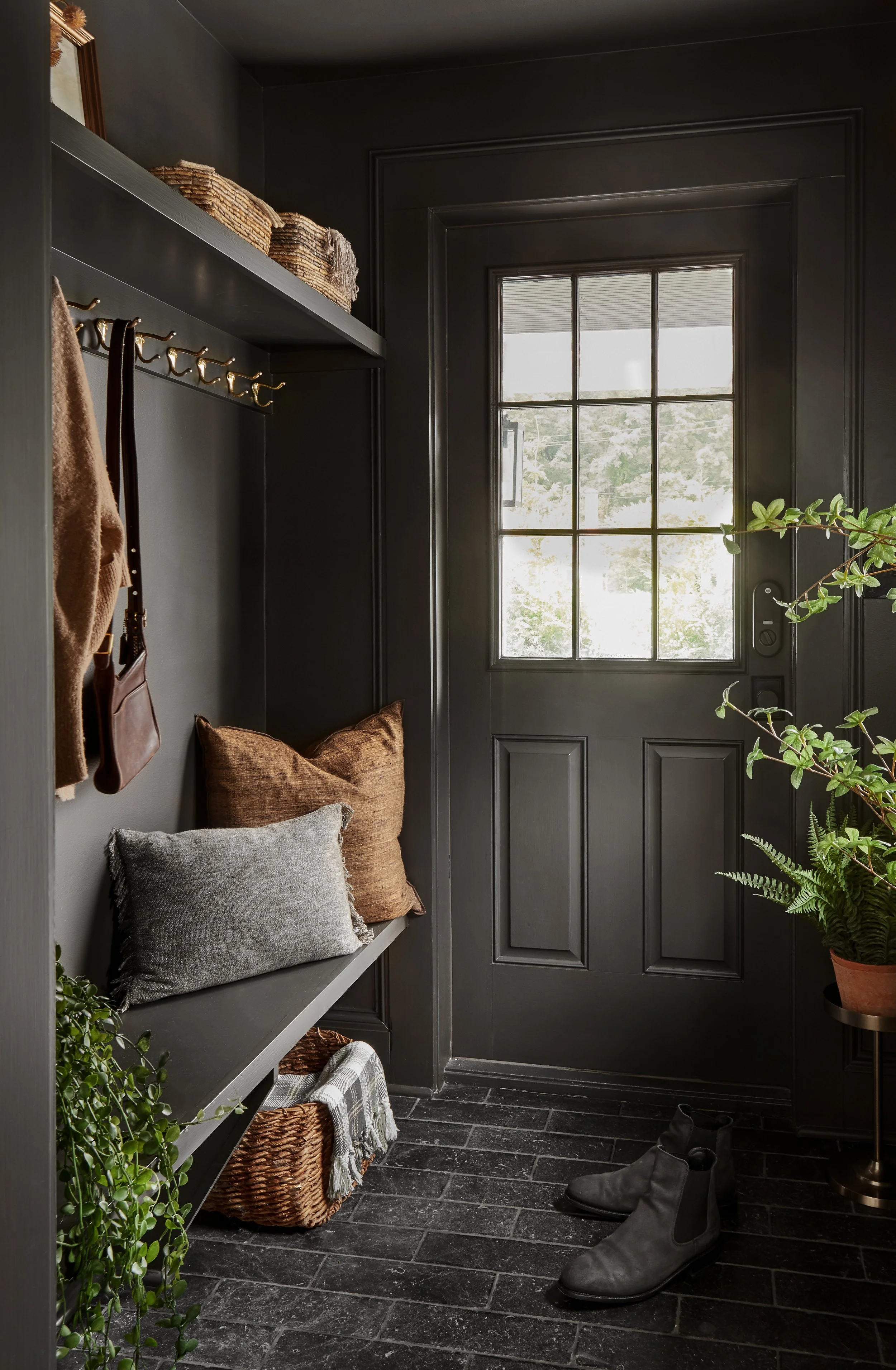 A dark-colored entryway with a windowed door, a black shelf with wicker baskets, hooks with coats and bags, pillows on a bench, a potted plant, and a pair of black boots on the floor.