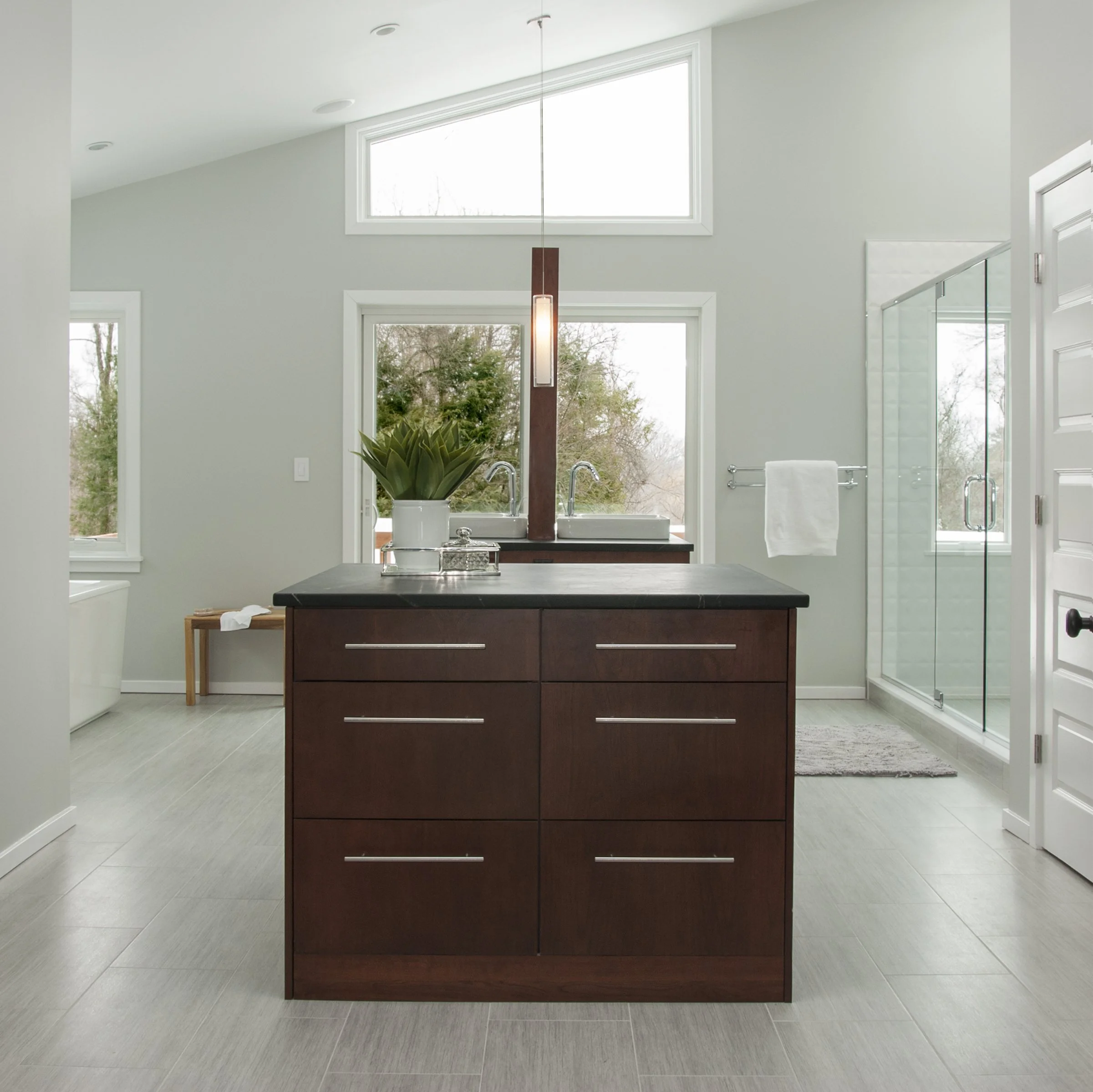Modern bathroom with a central dark wood vanity, double sinks, a glass shower stall, and large windows letting in natural light.