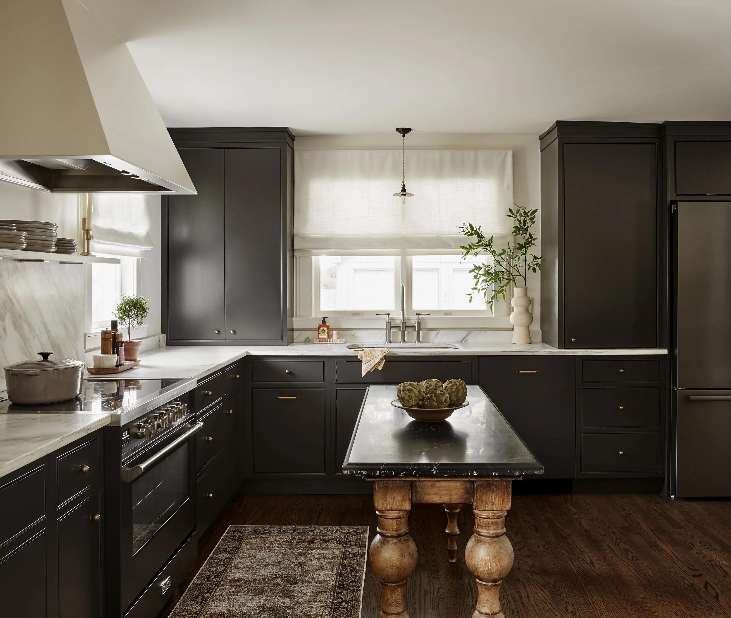 A modern kitchen with black cabinets, a marble countertop, a small island with a nero marble top, and hardwood floors. There are plants near the window.