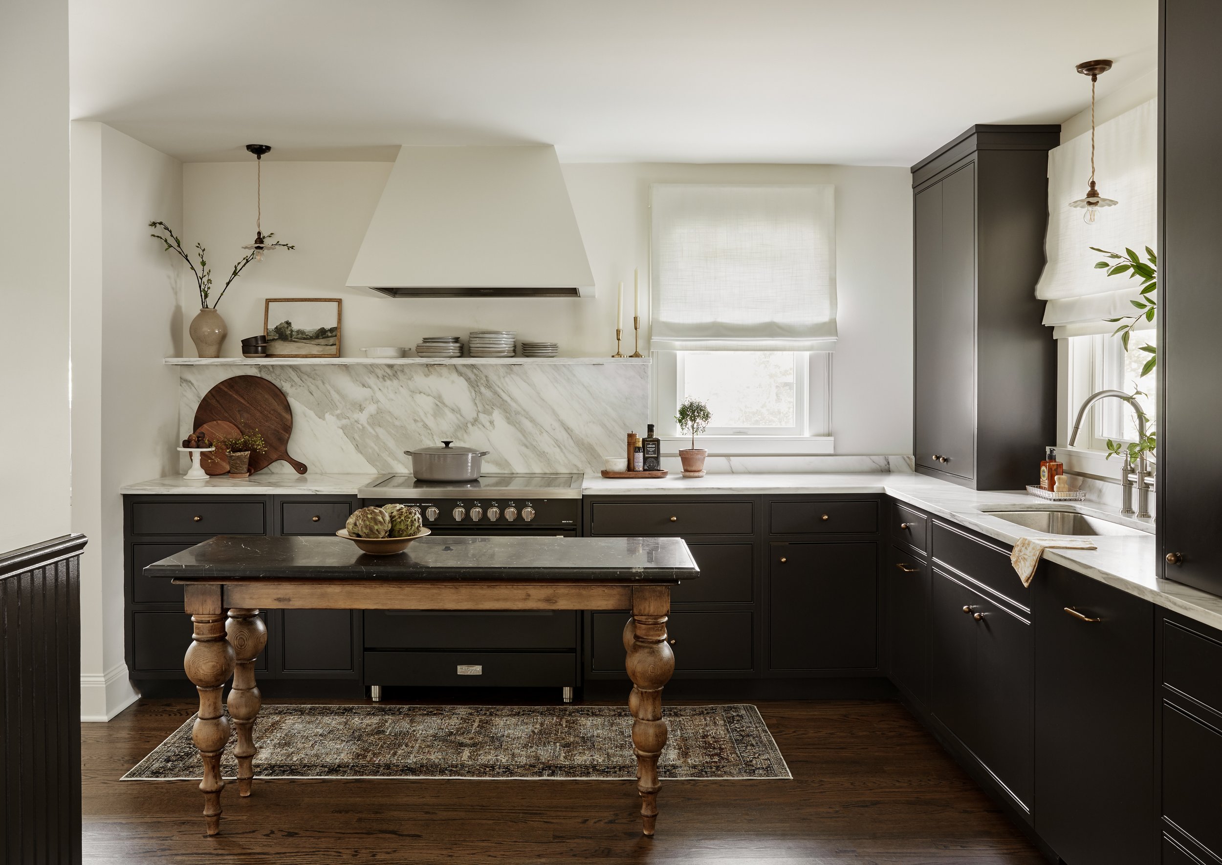 Modern kitchen with black cabinets, white marble countertops, a small kitchen island with turned wooden legs, dark wood flooring, a window with a White Roman shade, minimal decor including a vase with branches, a framed picture, and decorative plates, with a rustic rug on the floor.
