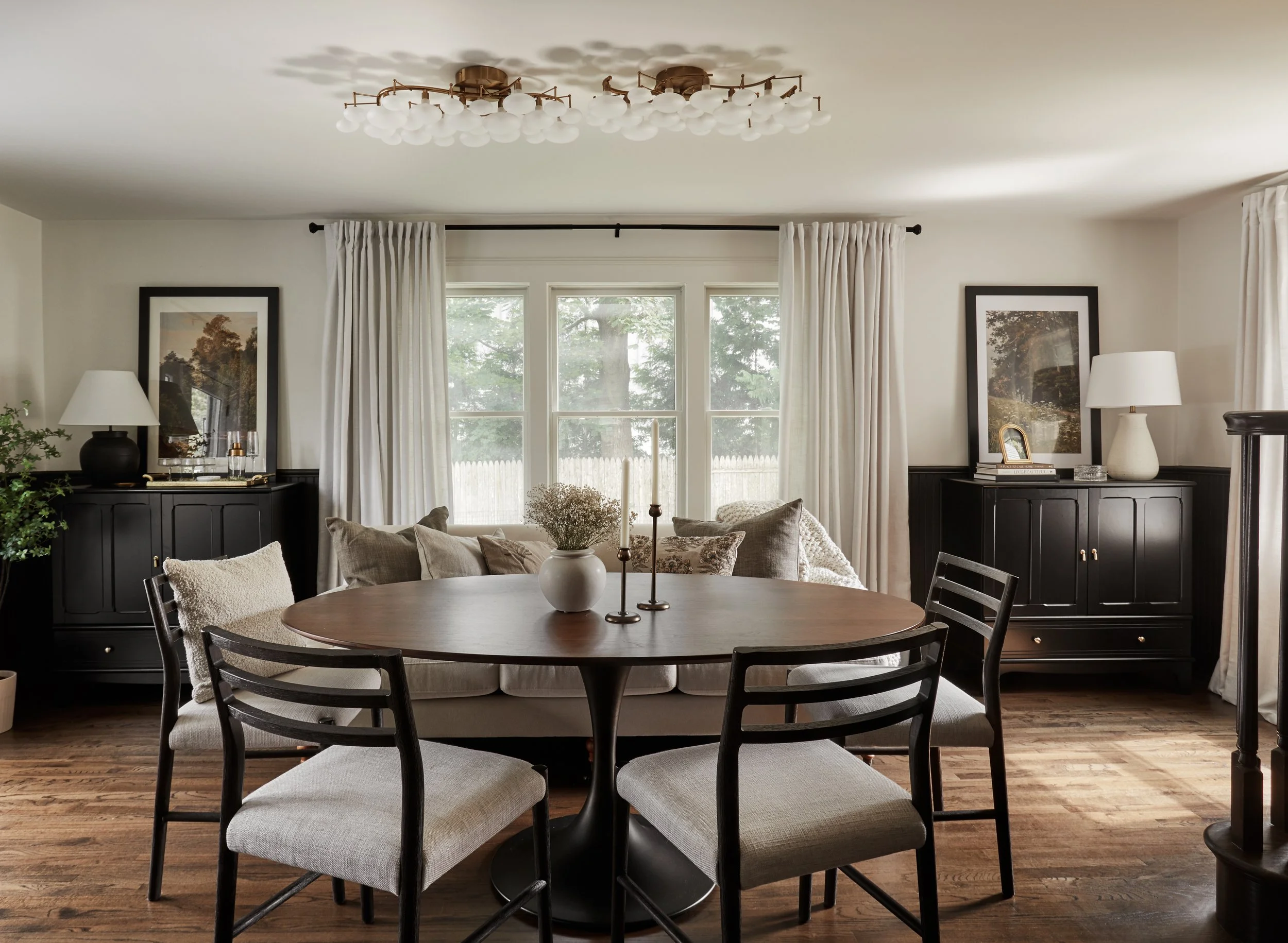 Living room with round dining table, black sideboards, lamps, framed pictures, white curtains, and a window with trees outside.