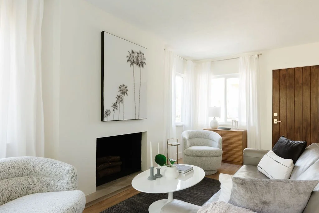 Living room with white walls, a white sofa with gray pillows, a round white coffee table, an armchair, a wooden sideboard, a lamp, and a large wall art of palm trees.