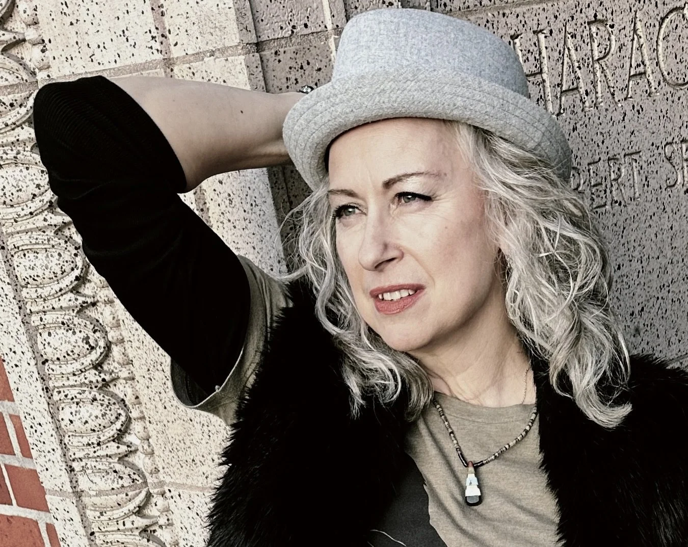 Margaret with gray curly hair wearing a light gray fedora hat, black top, and a necklace with a pendant, leaning against a brick wall with engraved words.