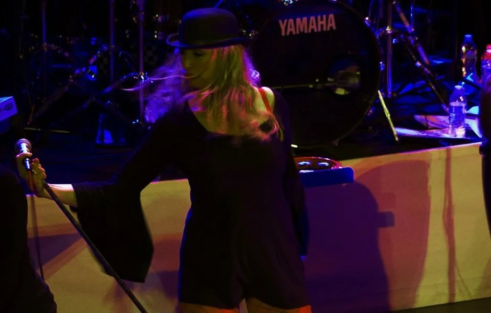 A woman in a black dress and hat holding a microphone on stage with a Yamaha drum set in the background, illuminated by colorful stage lights.