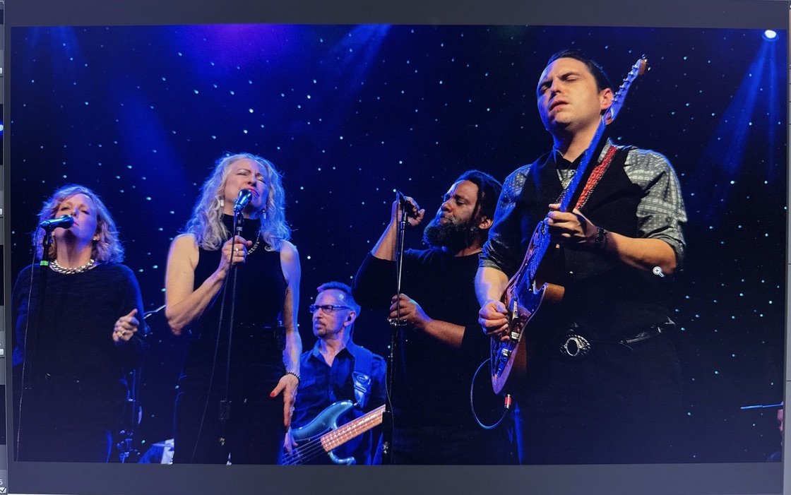 A band performs on stage with five members, three women and two men, singing and playing instruments, under a starry backdrop with blue stage lighting.