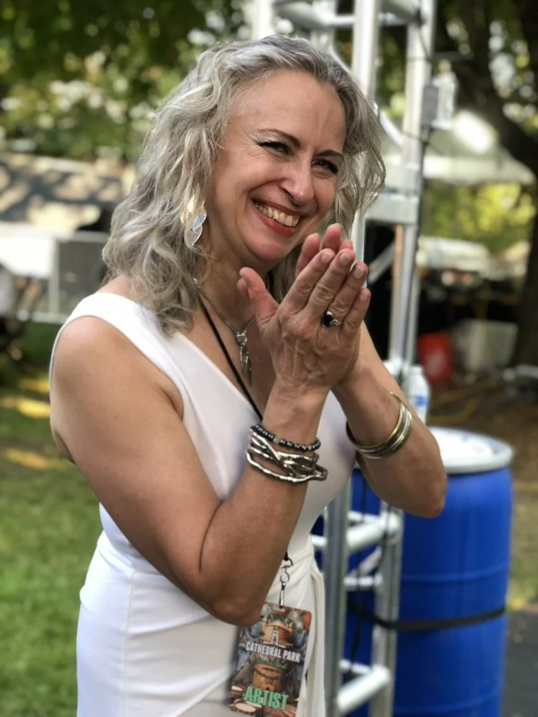 Margaret with curly gray hair smiling and clapping her hands outdoors, wearing a white sleeveless top and several bracelets, with a badge hanging around her neck. Background includes trees and event equipment.