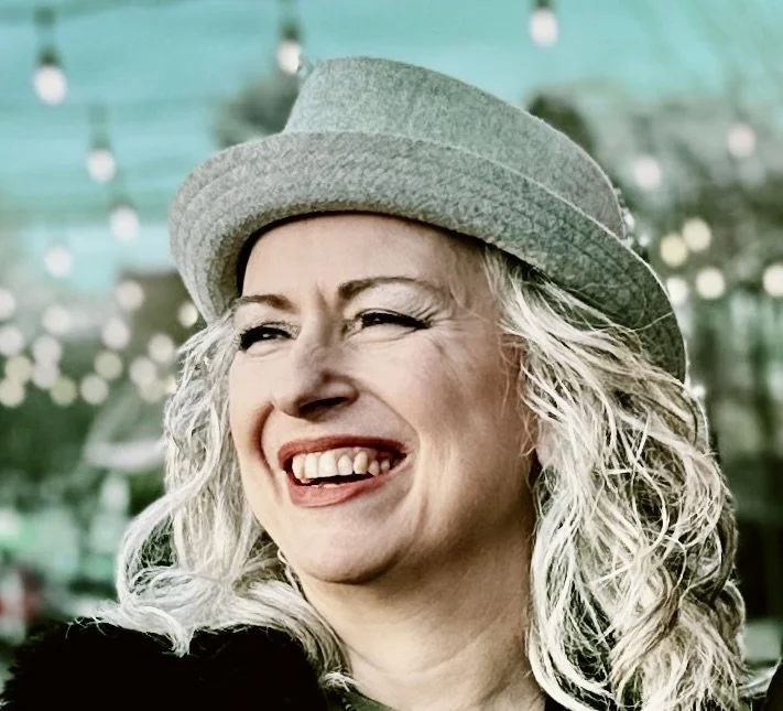A woman with curly gray hair wearing a gray hat, smiling broadly with missing front teeth, outdoors with blurred string lights in the background.
