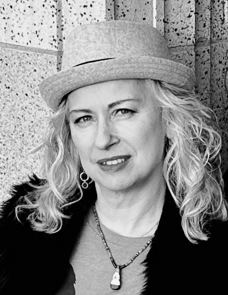 Black and white photo of Margaret with curly blonde hair wearing a light-colored hat, earrings, and a necklace, standing in front of a brick wall.