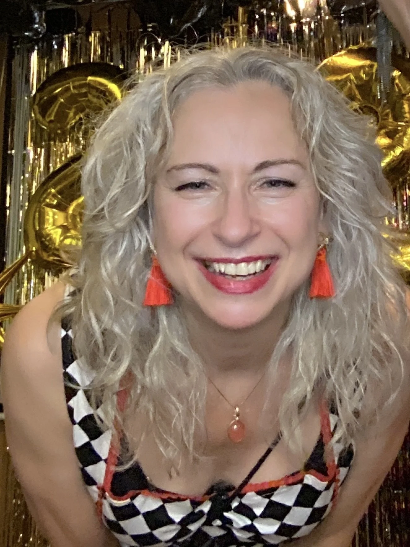 Margaret with curly blonde hair, wearing red tassel earrings and a checkered top, standing in front of gold balloons and tinsel decorations.
