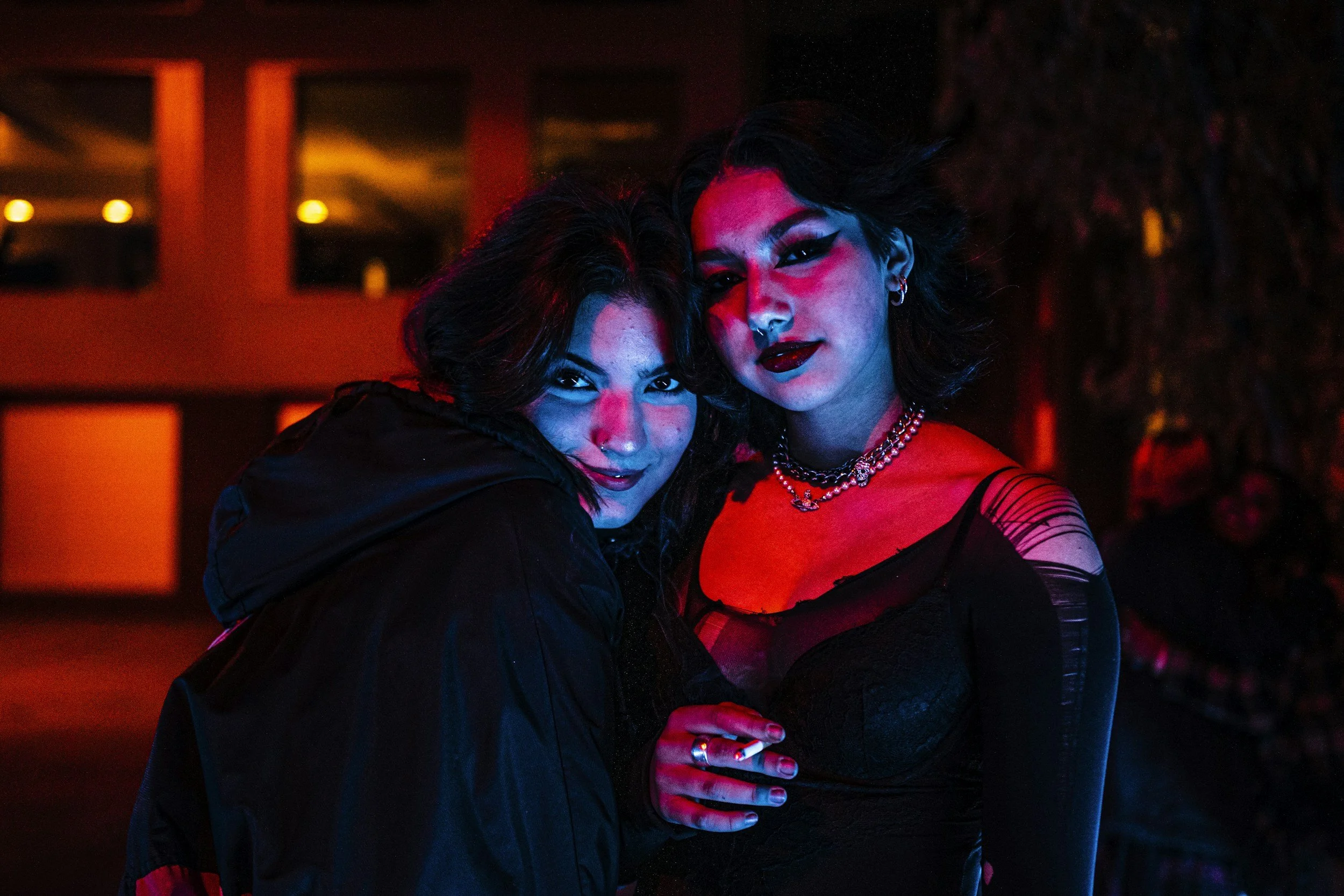 Two women with dark hair and makeup in a dimly lit, colorful nightclub or party setting, close together and looking at the camera.