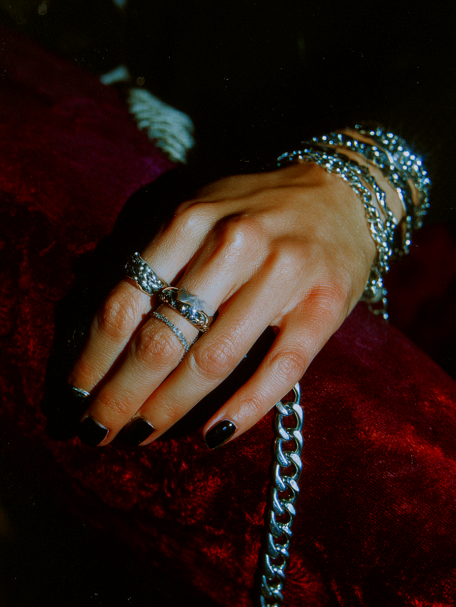 Close-up of a person’s hand resting on a red velvet surface, adorned with multiple silver rings, bracelets, and painted black nails.