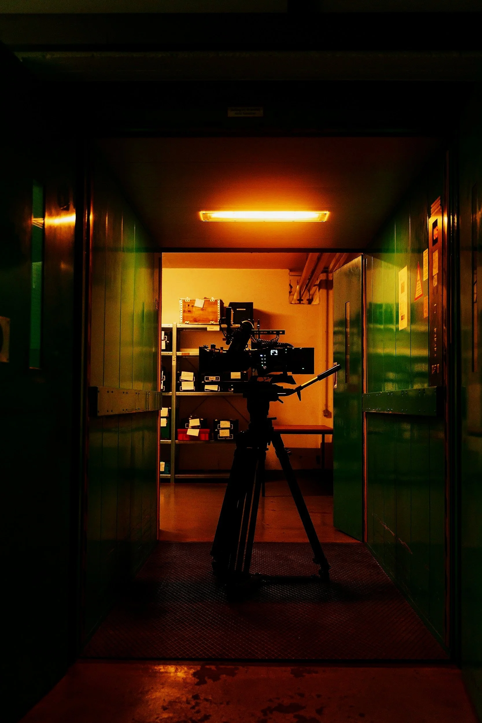 A silhouette of a camera mounted on a tripod inside a storage area or hallway, with shelves and boxes visible in the background, illuminated by a warm orange light overhead.