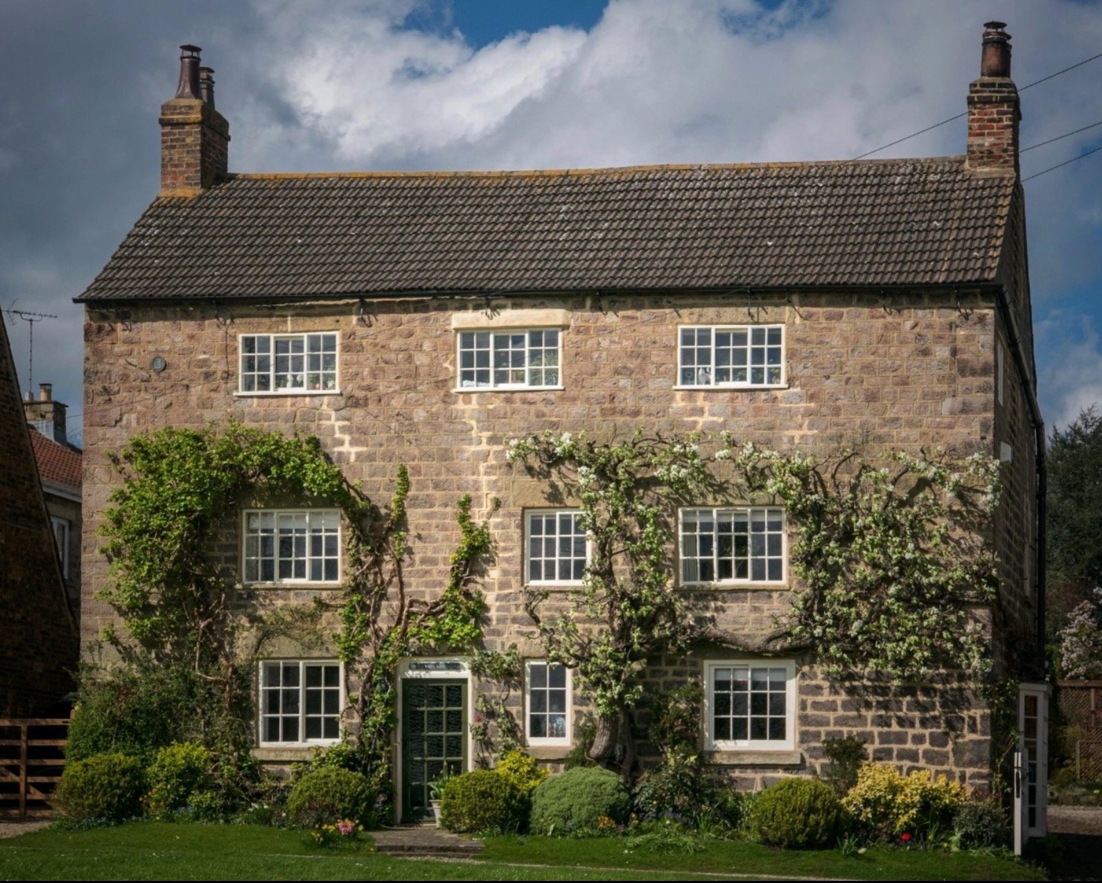 A stone house with a gabled roof, multiple centered windows, vines on the facade, and a well-maintained garden in front