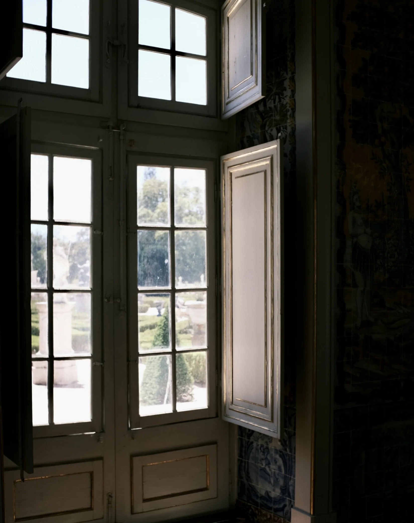 Open white window shutters on tall glass windows with a garden view outside.