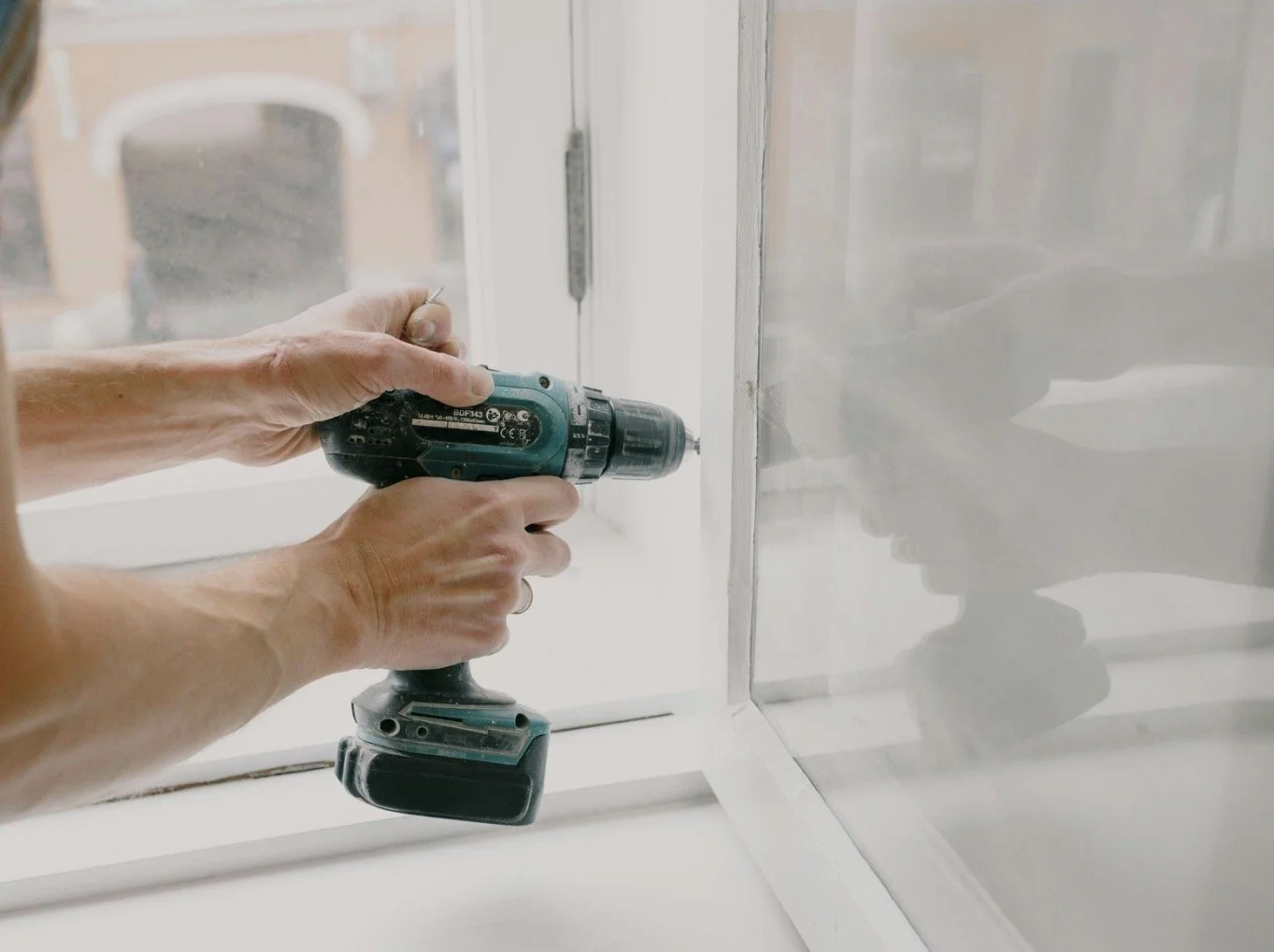 A person's hands using a cordless power drill to make a hole in a white wall or panel near a window.