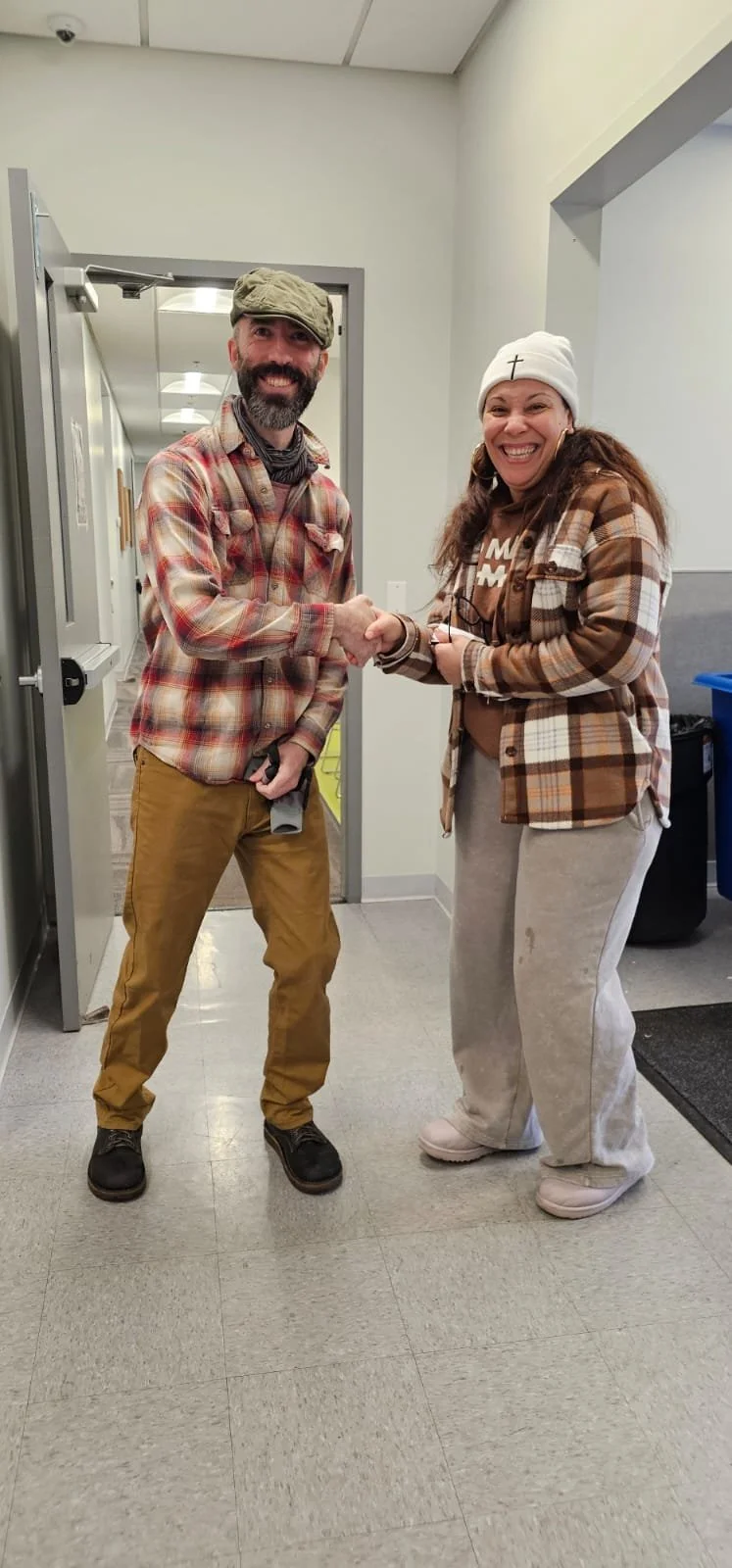 A man and woman smiling and shaking hands indoors, both wearing plaid shirts and casual clothing.